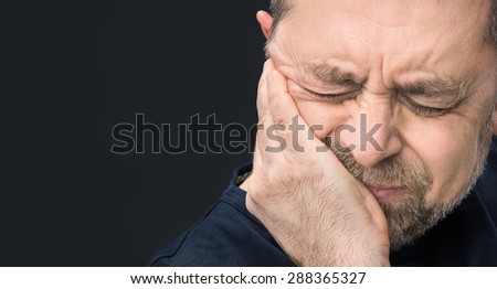 Headache. Portrait of an elderly man with face closed by hand on dark background with copy-space