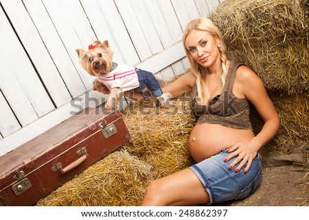 beautiful pregnant blonde woman with two braids sitting on hay with clothed dog near her