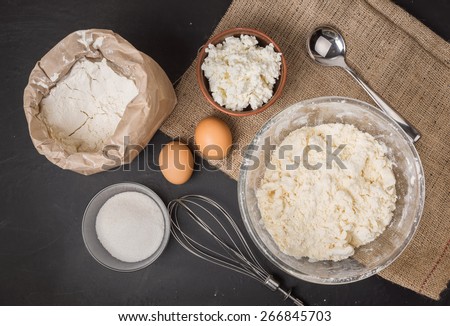 The ingredients for homemade cheesecake: curd in ceramic bowl, two eggs, sugar in a glass bowl, whisk,  wheat flour in a paper bag, iron spoon, wheat flour mixed with butter, on sacking, top view