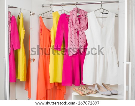 bright colorful female dresses hanging on coat hanger, pink scarf, shoes and handbag in white wardrobe