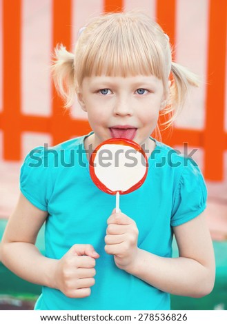 Funny child with candy lollipop, happy little blonde girl with blue eyes in a blue T-shirt eating big sugar lollipop, kid eat sweets in the park on the playground