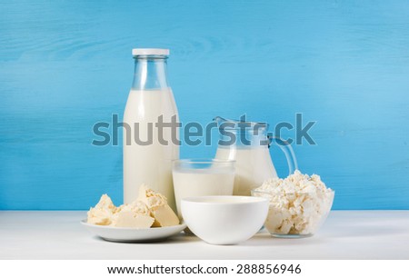 tasty healthy dairy products on a white table on a blue background: sour cream in a white bowl, cottage cheese in bowl,  butter on a saucer, milk in a jar, glass bottle and in a glass