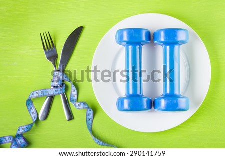 ?oncept of a healthy lifestyle, diet, sports, weight loss, anti-obesity, exercise, healthy diet.   Dumbbells on a plate, crossed fork and knife wrapped centimeter on a green table top view, closeup