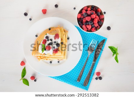 Delicious sweet crepes decorated air cream and ripe berries, raspberries, blackberries and blueberries on a white plate, fork, knife, bright blue polka dot napkin. Top view