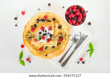 Delicious sweet crepes decorated air cream and ripe berries, raspberries, blackberries and blueberries on a white plate, fork, knife, white napkin. Top view
