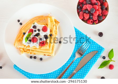Delicious sweet crepes decorated air cream and ripe berries, raspberries, blackberries and blueberries on a white plate, fork, knife, bright blue polka dot napkin. Top view