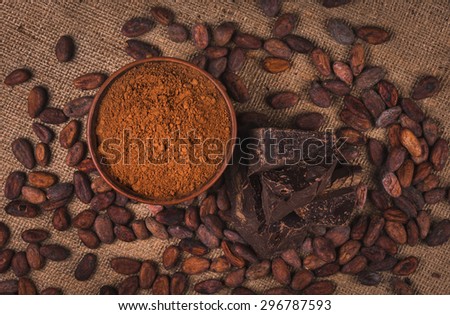 crude dark cocoa powder in a brown ceramic bowl, raw cocoa beans in the peel and raw chocolate on sacking close up, top view, ingredients for preparing chocolate and sweets