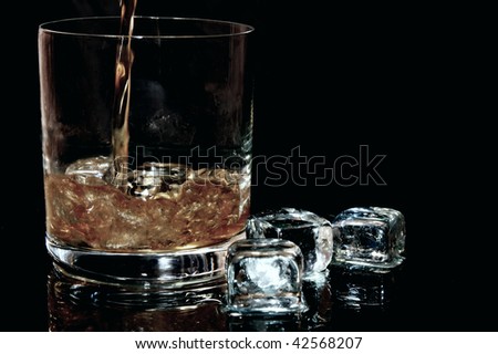Whiskey flows into a glass with ice cubes to the left against black background in the horizontal format