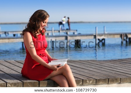 Brunette woman dress in red reading a book on the lake jetty