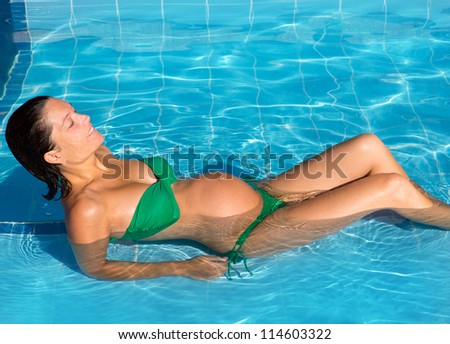 Beautiful pregnant woman sun tanning relaxed at blue pool with green bikini
