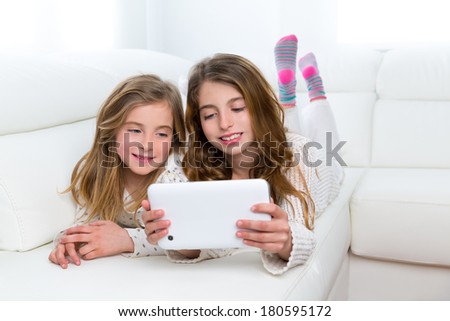 Children sister friends kid girls playing together with tablet pc lying on white sofa
