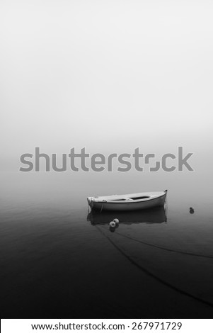 Small boat without oars tied with ropes floating in the sea in black and white during a foggy sunrise
