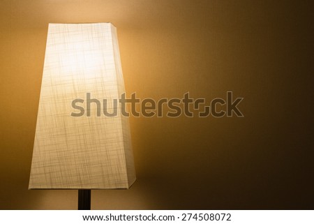 A lamp with a cloth lamp shade in a dark room against a simple wall.