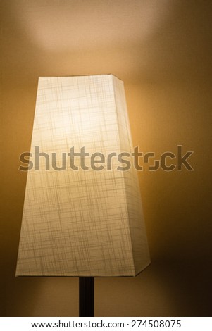 A lamp with a cloth lamp shade in a dark room against a simple wall.