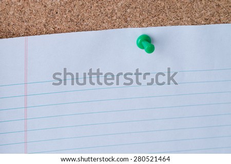 A green thumb tack holding a white piece of lined loose leaf paper on a cork board.