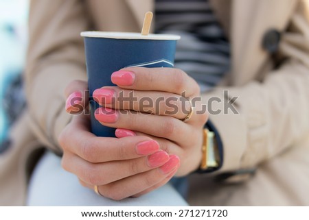 Blue paper Cup of coffee in female hands with pink manicure. The girl is dressed in a beige coat, striped t-shirt and blue jeans.