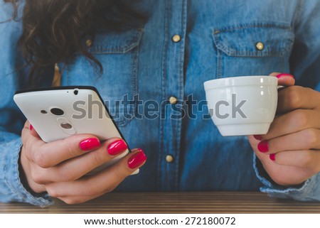 The girl in the blue denim shirt sitting at the table. In one hand a white Cup of espresso, and the other with a white mobile phone. Red manicure. Vintage photo.