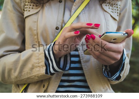 The girl uses the phone close up, she is dressed in a beige jacket and striped shirt, at the shoulder yellow strap from the bag. She holds a white phone with two hands with red nail Polish.