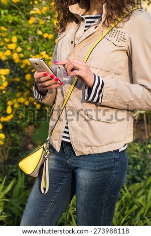 Girl uses the phone on the background of yellow flowers, she is dressed in a beige jacket, blue jeans and a striped shirt, at the shoulder yellow gray bag. In her hand with red manicure white phone.