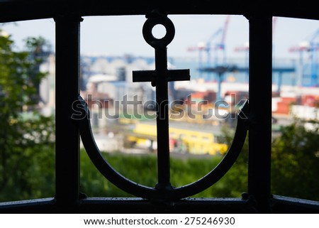 In the foreground closeup you can see the silhouette of an anchor in a metal fence. On the back you can see the blurry term commercial sea port and cranes.