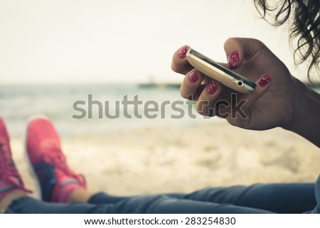 Girl resting on the beach and using a mobile phone. A young girl dressed in jeans and pink sneakers.