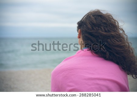 Young woman with curly hair sitting alone on the beach in cloudy weather and looks out to sea. Her hair windblown. On her shoulders is pink plaid. Pensive mood.