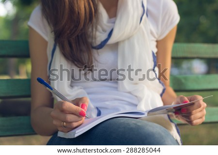 Young female student sitting on a bench in the park. She takes notes in a notebook. She is dressed in blue jeans and white T-shirt. Close-up.
