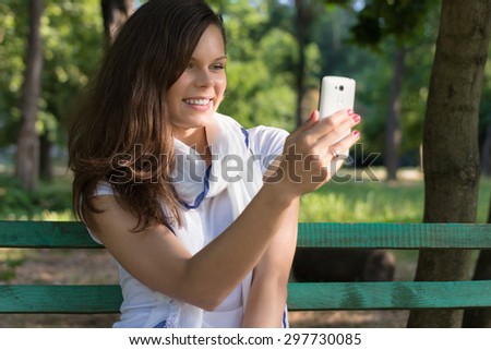 Young happy smiling woman sitting on a Park bench in the shade of the trees and makes a picture on her smart phone. Holiday summer in the Park on a Sunny day.