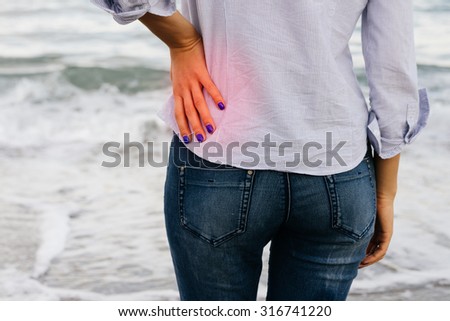 Low Back Pain. The woman in jeans and shirt standing on the shore and holding her lower back.
