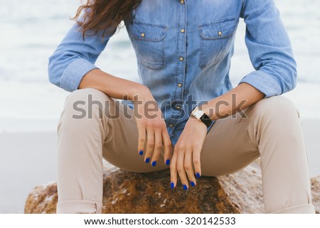Woman in beige pants and a denim shirt sitting on a rock by the sea. Shirt sleeves rolled up, watch on her arm, a blue manicure.