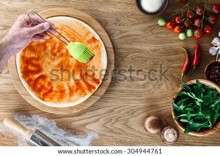 Pizza cooking process: spreading tomato sauce on pizza base with a silicone pastry brush. Wooden background, top view.