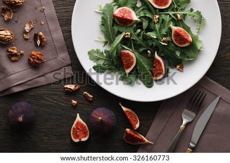 A salad with green leaves of rocket and spinach, fig pieces and walnuts in a white plate on dark wooden background. Top view.