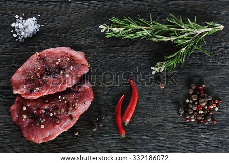 Two beef steaks garnished with a couple of rosemary twigs, dwarf chili peppers, sea salt granules and peppercorns on a dark background. Top view.