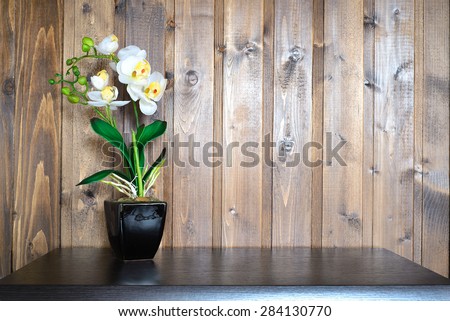 Artificial flower in a vase standing on a wooden pedestal on a background of a wooden wall