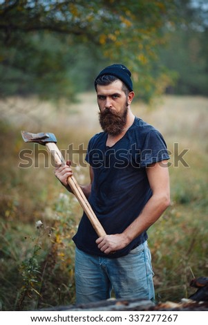 brutal bearded lumberjack in a warm cap and a T-shirt holding an ax in the woods on a background of trees