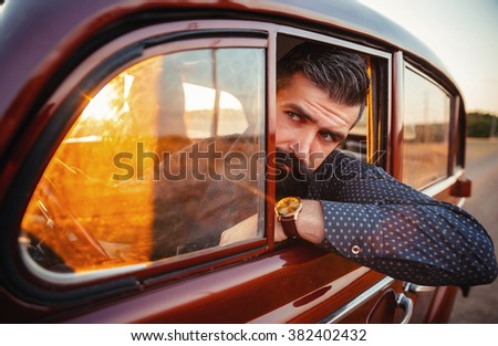 brutal bearded man with a mustache in a shirt, pants with suspenders with a girl with dark hair and big lips with bright red lipstick in a short dress and heels near retro car at sunset