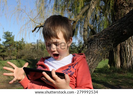 The boy looks at the plate and his face surprised.  The child is in the nature, on the background of trees and sky. One hand holds a tablet, on the other hand, the fingers spread wide apart.