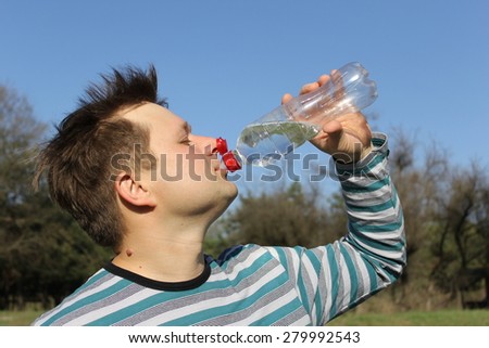 A man drinks water from a plastic bottle. In the background, the sky and the green trees. The man\'s face in the foreground. The person and a bottle of focus. Hand of man holding a bottle in his mouth.