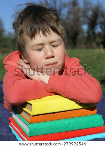The boy is lying on a pile of books. His hands crossed on books, his face is on the hands. His eyes are closed, he is sleeping. In the background blue sky and green trees. Books have colorful covers.