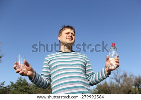 A man holding a plastic bottle of water and a glass. The background of blue sky and green trees. Man holding hands to the sides. His eyes are closed. He is happy and relaxed.