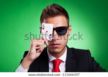 Young business man with sunglasses covers one eye with an ace of hearts. Over green background.