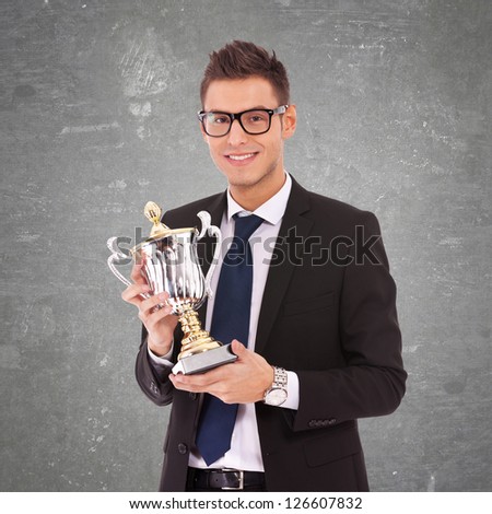 young business man  with glasses holding a big trophy on gray background