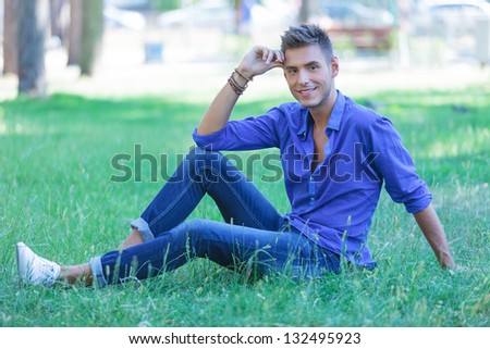 young casual man posing in the grass in a seated pose, smiling at the camera