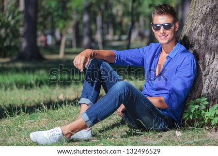 young casual man sitting on the ground and leaning to a tree trunk while smiling at the camera