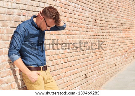 casual young man standing with a hand in his pocket and the other at the back of his head while looking down with melancholy, next to a brick wall