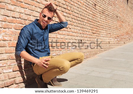 casual young man goofing around while sitting crouched by a brick wall, scratching his head and pulling his tongue out, while looking at the camera
