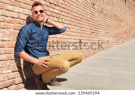 casual young man crouching next to a brick wall and holding a hand at the back of his neck while smiling to the camera