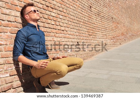 casual young man sitting in a crouched position with his back by a brick wall and looking away from the camera