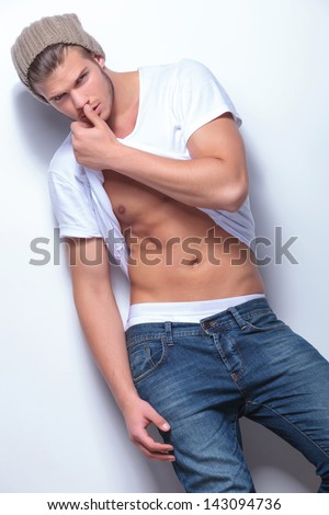 Young Fashion Man Pulling His T-Shirt Up And Looking At The Camera While Touching His Nose With His Thumb. On Light Gray Background