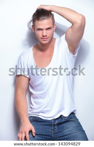 young fashion man holding back his hair while leaning with his back on a studio wall. on light gray background